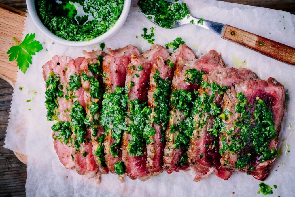 Sliced grilled barbecue beef steak with green chimichurri sauce on wooden background
