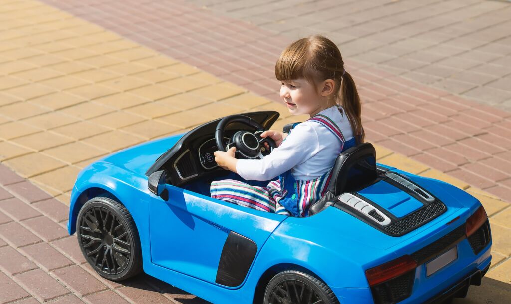 A Little Girl Is Driving A Battery-Powered Car In An Open-Air Amusement Park. A cute child is sitting in his blue toy car. The concept of a happy childhood. Entertainment for children on weekends.