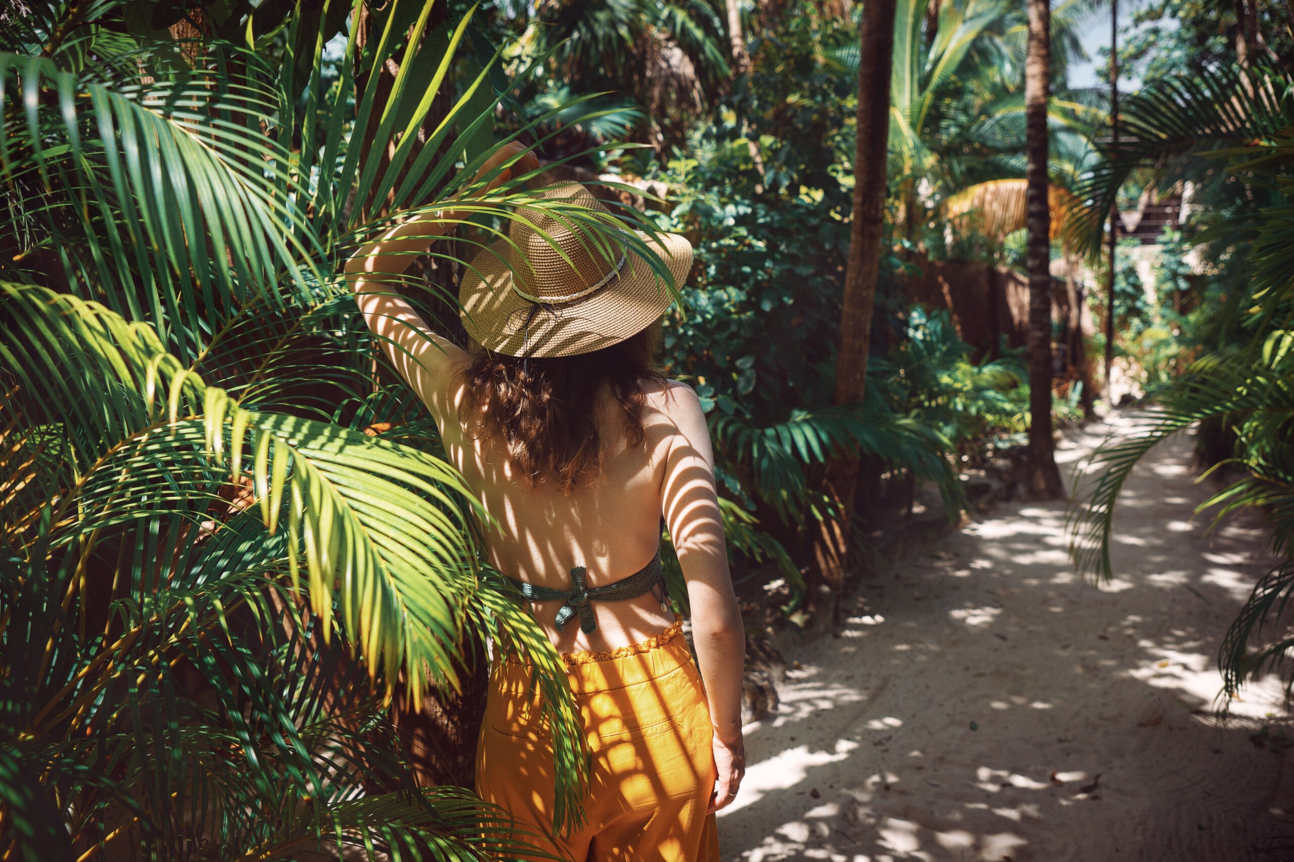 Young beautiful woman stands in the shade of palm trees, view from the back. A girl walks among tropical greenery on a bright sunny day in Mexico, Tulum