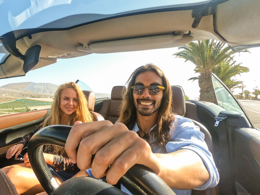 Happy young couple having fun during road trip in convertible car. Romantic lovers enjoying time together driving cabriolet auto in luxury vacation in a tropical city. Holiday and relationship concept