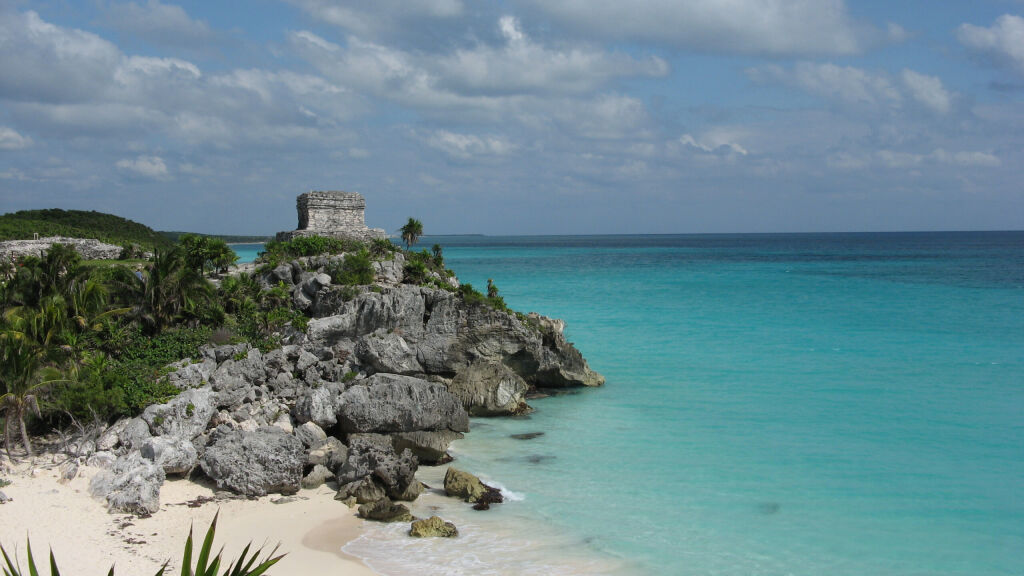 Mexico, Yucatan, Tulum Ruins - ancient Maya Temple of the god of winds on the beach, with clear turquoise water and cloudy sky rocks and vegetation, seen from afar 